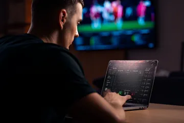 Um homem comemorando uma vitória em seu laptop enquanto símbolos de jogos flutuam ao redor, capturando a emoção dos jogos QWE123.